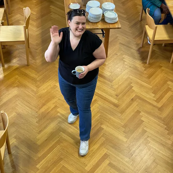 Eine Frau in einem schwarzen T-Shirt und Jeans winkt, während sie eine Kaffeetasse in einem Raum mit Holzstühlen und einem Tisch mit Tellern hält.