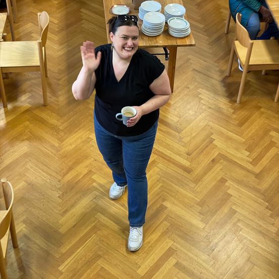 Eine Frau in einem schwarzen T-Shirt und Jeans winkt, während sie eine Kaffeetasse in einem Raum mit Holzstühlen und einem Tisch mit Tellern hält.