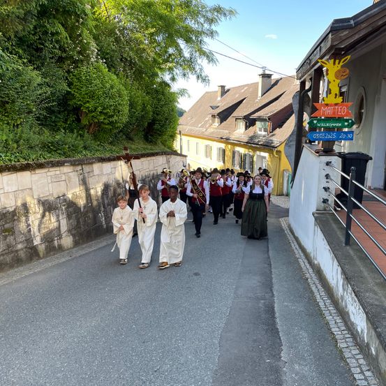 Eine Gruppe von Menschen in weißen Gewändern zieht in einer Prozession die Straße hinunter und trägt ein Kreuz. Ihnen folgt eine Band in traditioneller Kleidung.