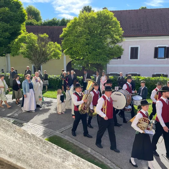 Eine Blaskapelle in traditioneller Kleidung spielt im Freien, mit Zuschauern und einem Gebäude im Hintergrund.