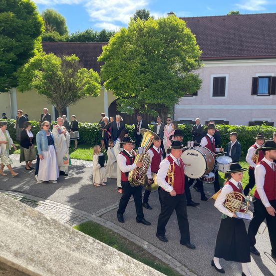 Eine Blaskapelle in traditioneller Kleidung spielt im Freien, mit Zuschauern und einem Gebäude im Hintergrund.