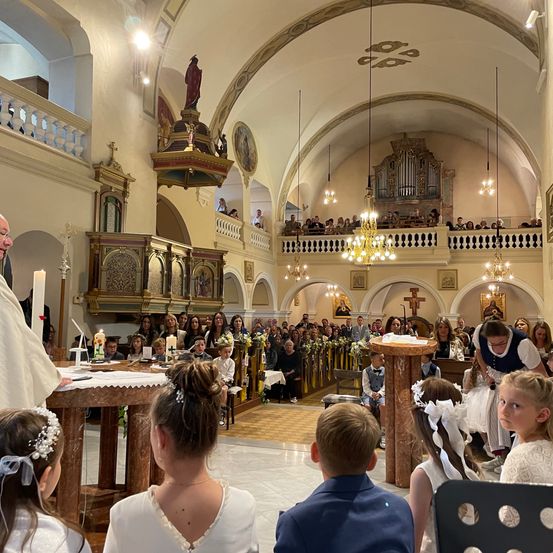 Ein Priester leitet eine religiöse Zeremonie in einer Kirche. Altäre mit Kerzen und Blumen sind aufgestellt. Kinder in weißen Kleidern sitzen vorne, und die Gemeinde steht dahinter.