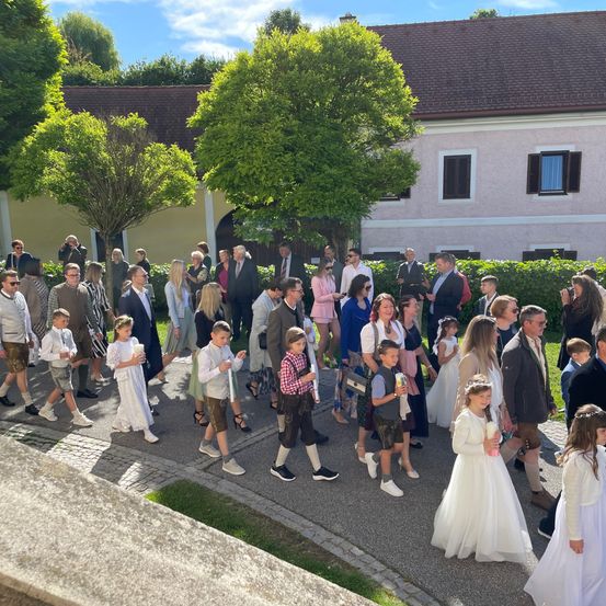 Eine große Gruppe von Menschen, darunter Kinder in traditioneller Kleidung, geht in einer Reihe vor einem Gebäude bei teilweise bewölktem Himmel.