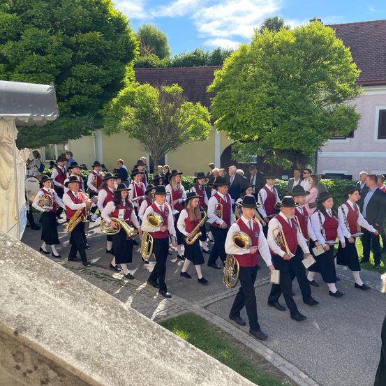 Eine Blaskapelle, gekleidet in rote Westen und schwarze Hüte, spielt Instrumente, während sie einen von Bäumen und einem Gebäude gesäumten Weg entlanggehen. Zuschauer beobachten sie.