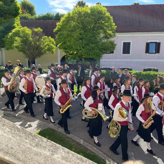 Eine Blaskapelle in roten Westen und schwarzen Röcken geht auf einem gepflasterten Weg und spielt verschiedene Instrumente. Hinter ihnen steht ein Haus mit Bäumen und Sträuchern.