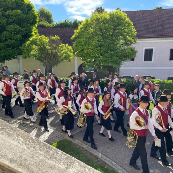 Eine Blaskapelle in roten Westen und schwarzen Hosen tritt im Freien auf, wobei Zuschauer dahinter zusehen. Die Band spielt verschiedene Instrumente, darunter Saxophone, Trompeten und Tuben.