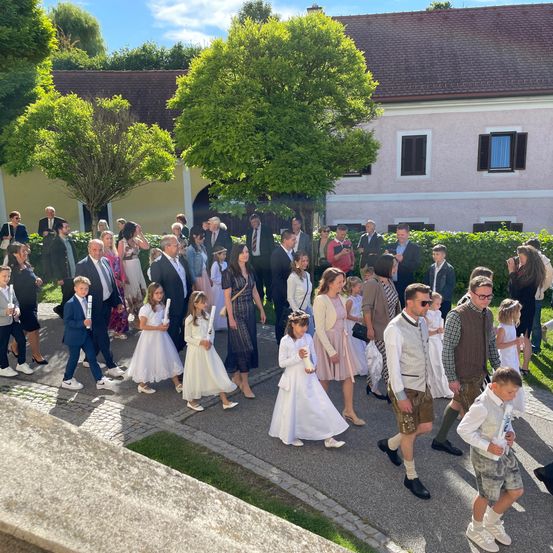 Eine große Gruppe von Menschen, darunter Kinder in weißen Kleidern und Jungen in Anzügen, geht vor einem Gebäude mit Bäumen und Büschen.