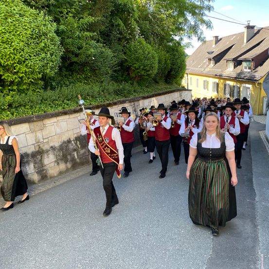Eine Musikkapelle, gekleidet in rote Westen, tritt auf der Straße auf. Hinter ihnen beobachtet eine Frau in traditioneller Tracht. Im Hintergrund befindet sich ein gelbes Gebäude.