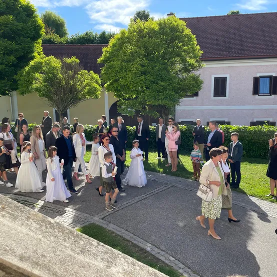 Eine Gruppe von Menschen, darunter Kinder in weißen Kleidern, geht auf einem Pfad vor einem Gebäude. Einige halten Kerzen, während andere auf dem Rasen oder der Veranda stehen. Bäume und Büsche sind in der Nähe.