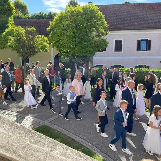 Eine Gruppe von Menschen, darunter Kinder in weißen Kleidern und Männer in Anzügen, gehen auf einem Pfad vor einem Gebäude mit braunem Dach und weißen Wänden.