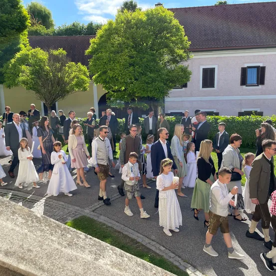 Eine große Gruppe von Menschen, die in Weiß gekleidet sind, zieht in einer Prozession auf einer Straße vor einem Gebäude. Einige tragen traditionelle bayerische Kleidung. Bäume und Sträucher sind in der Umgebung gepflanzt.