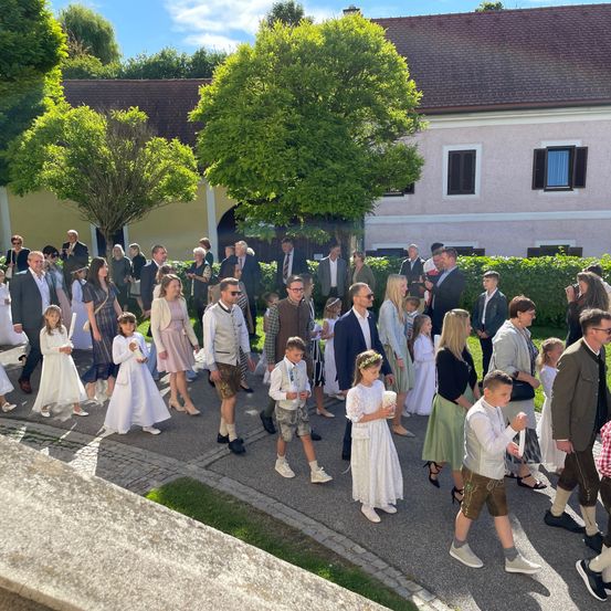 Eine große Gruppe von Menschen, die in Weiß gekleidet sind, zieht in einer Prozession auf einer Straße vor einem Gebäude. Einige tragen traditionelle bayerische Kleidung. Bäume und Sträucher sind in der Umgebung gepflanzt.