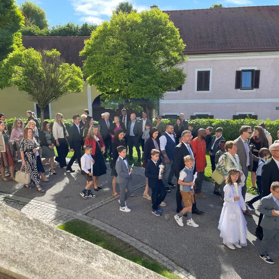 Eine Gruppe von Menschen, einschließlich Kindern, ist vor einem Gebäude versammelt. Sie sind in verschiedenen formellen und lässigen Kleidungsstücken gekleidet und einige halten Kerzen. Das Gebiet ist von Bäumen und Büschen umgeben.