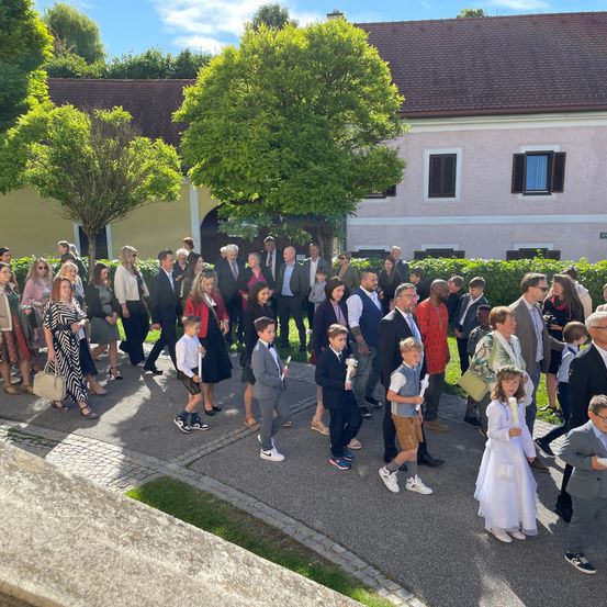 Eine Gruppe von Menschen, einschließlich Kindern, ist vor einem Gebäude versammelt. Sie sind in verschiedenen formellen und lässigen Kleidungsstücken gekleidet und einige halten Kerzen. Das Gebiet ist von Bäumen und Büschen umgeben.