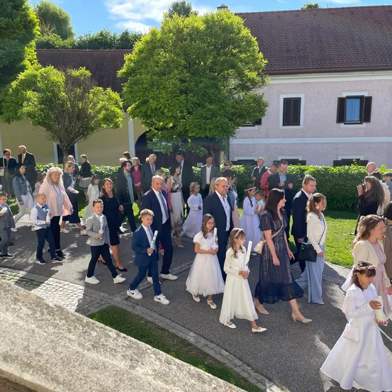Eine Gruppe von Menschen, einschließlich Kindern, geht in einer Reihe vor einem Gebäude. Sie tragen formelle Kleidung und halten Kerzen. Das Gebäude hat ein braunes Dach und weiße Wände mit schwarzen Fensterläden.