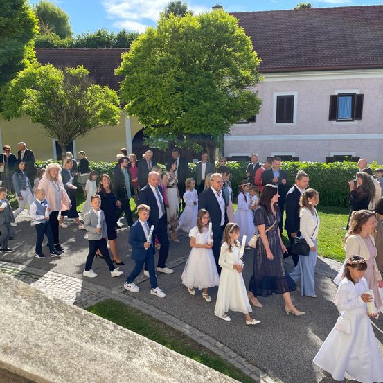 Eine Gruppe von Menschen, einschließlich Kindern, geht in einer Reihe vor einem Gebäude. Sie tragen formelle Kleidung und halten Kerzen. Das Gebäude hat ein braunes Dach und weiße Wände mit schwarzen Fensterläden.