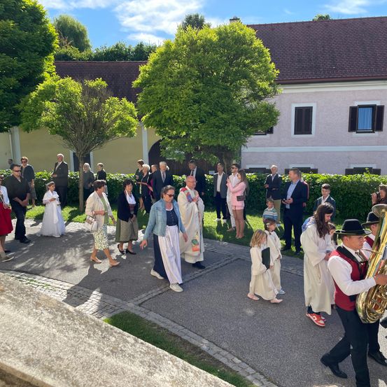 Eine Gruppe von Menschen in traditioneller Kleidung, einschließlich Kinder, geht an einem sonnigen Tag vor einem Gebäude. Einige tragen Bücher.