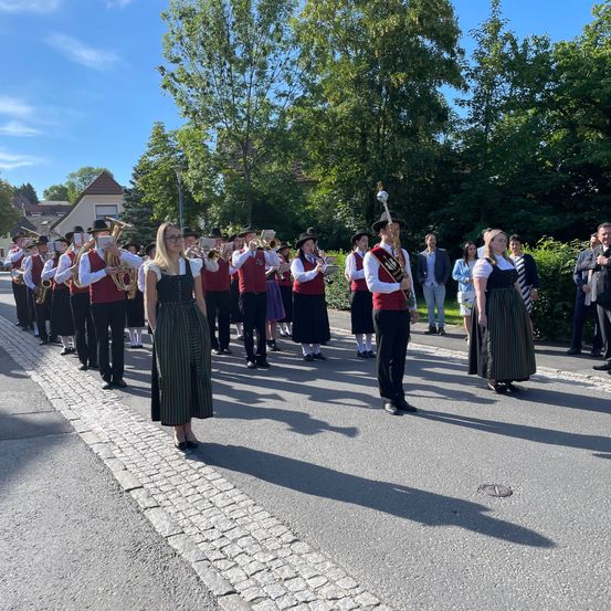 Eine Blaskapelle in traditioneller Kleidung spielt auf der Straße, während Zuschauer an der Seite stehen.