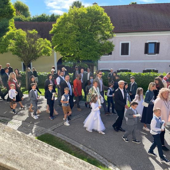Eine große Gruppe von Menschen in formeller Kleidung, darunter Kinder, gehen in einer Prozession vor einem Gebäude. Das Gebäude hat ein braunes Dach und mehrere Fenster. Der Boden ist mit Kopfsteinen gepflastert, und es gibt Bäume und Büsche in der Umgebung.