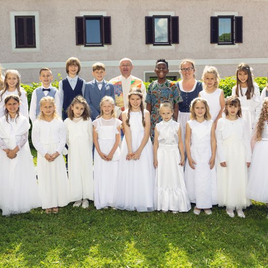 Eine Gruppe von Kindern in weißen Kleidern und Jungen in Anzügen steht vor einem Gebäude. Einige halten Händchen, und Erwachsene sind ebenfalls anwesend.