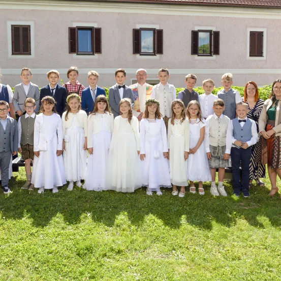 Eine Gruppe von Kindern und Erwachsenen steht vor einem Gebäude. Die Kinder sind in Weiß gekleidet, und zwei Erwachsene tragen formelle Kleidung. Sie scheinen einen besonderen Anlass zu feiern.