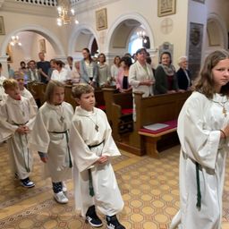 Kinder in weißen Gewändern stehen in einer Kirche, mit Erwachsenen im Hintergrund und Bänken im Vordergrund.