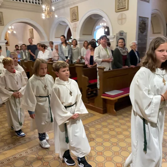 Kinder in weißen Gewändern stehen in einer Kirche, mit Erwachsenen im Hintergrund und Bänken im Vordergrund.