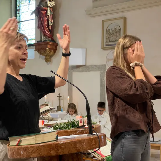 Eine Frau steht in einer Kirche an einem Podium, ihre Hände erhoben, während sie singt. Eine andere Frau, die ihr Gesicht bedeckt, steht in der Nähe. Eine Statue und ein gerahmtes Bild schmücken die Wand hinter ihnen.