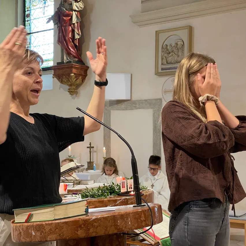 Eine Frau steht in einer Kirche an einem Podium, ihre Hände erhoben, während sie singt. Eine andere Frau, die ihr Gesicht bedeckt, steht in der Nähe. Eine Statue und ein gerahmtes Bild schmücken die Wand hinter ihnen.
