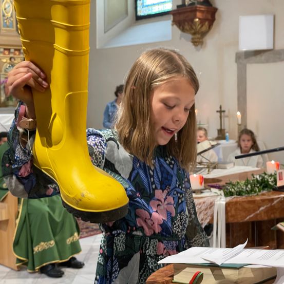 Ein Mädchen hält einen gelben Stiefel und schaut nach oben, in einer Kirche mit Menschen und Kerzen.