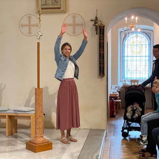 Eine Frau in einer Kirche winkt mit beiden Händen. Ein Mann steht hinter einem Kinderwagen mit einem Baby. Ein Feuerlöscher ist in der Nähe.