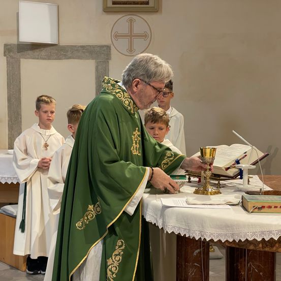 Ein Priester in einer grünen Gewandung steht am Altar, liest aus einem Buch, mit einem goldenen Kelch und Messdienern in der Nähe.
