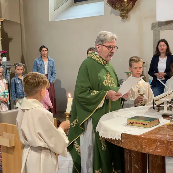 Ein Priester steht in einer Kirche an einem Tisch und liest von einem Papier. Ein Junge hält eine Kerze. Hinter ihnen stehen zwei weitere Jungen und mehrere Frauen.