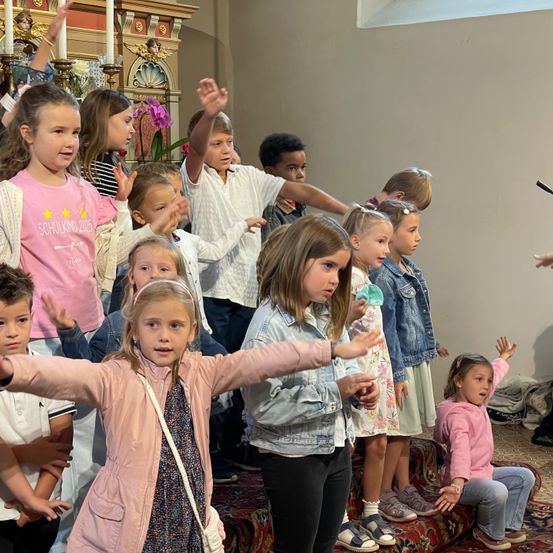 Eine Gruppe von Kindern steht in einer Kirche, einige heben die Hände. Ein Mädchen in Pink steht vorne und in der Mitte, während andere Jacken und Turnschuhe tragen.