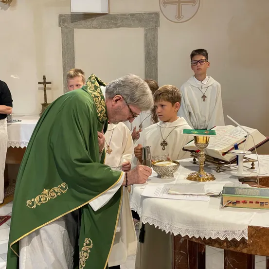 Ein Priester in grünen Gewändern wäscht die Füße eines jungen Jungen, mit anderen Kindern und einem Kreuz im Hintergrund.
