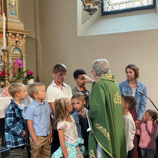 Ein Priester segnet eine Gruppe von Kindern während eines Gottesdienstes. Die Kinder stehen in einer Reihe und hören aufmerksam zu.