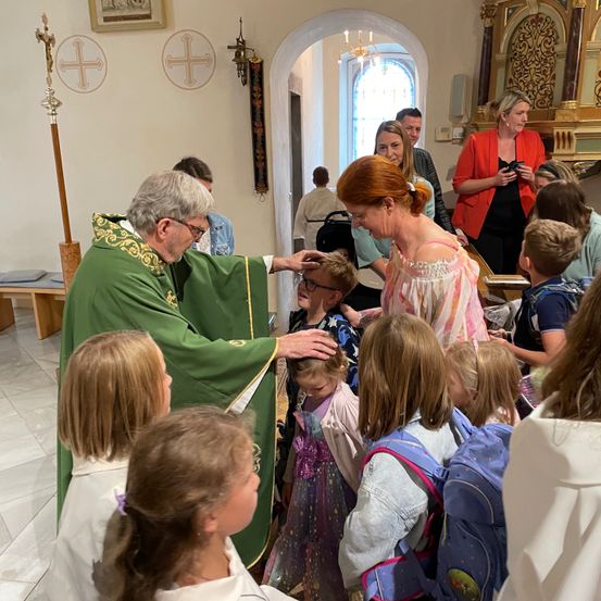 Ein Priester segnet Kinder in einer Kirche. Eine Frau in Pink hält ein kleines Kind. Andere Kinder sind um sie herum versammelt.