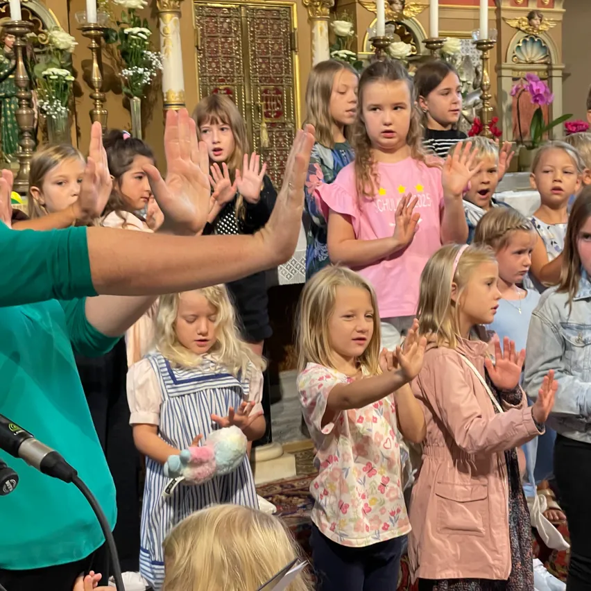 Eine Gruppe junger Mädchen und ein Mann befinden sich in einer Kirche. Die Mädchen singen, und einige heben ihre Hände. Der Mann hält ein Mikrofon. Der Altar hat Kerzen, Blumen und Statuen.