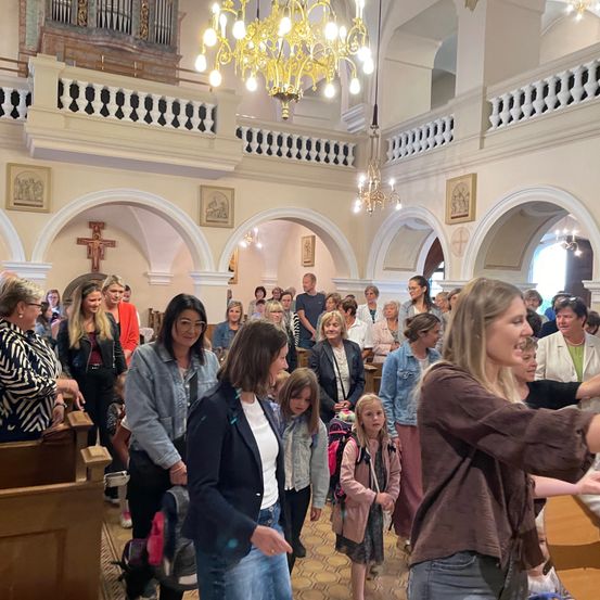 Eine Gruppe von Menschen, darunter Frauen und Kinder, steht in einer Kirche. Einige tragen Rucksäcke. Die Kirche hat Kronleuchter und gerahmte Bilder an den Wänden.