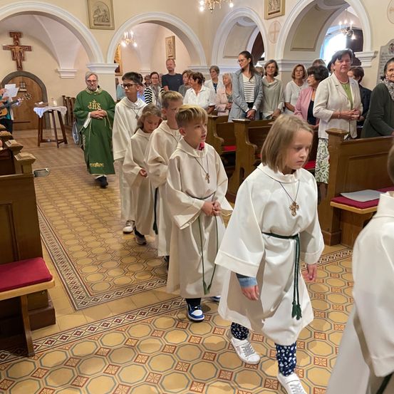 Eine Gruppe von Kindern in weißen Gewändern und Turnschuhen geht während einer Zeremonie in einer Kirche, begleitet von Erwachsenen im Hintergrund.