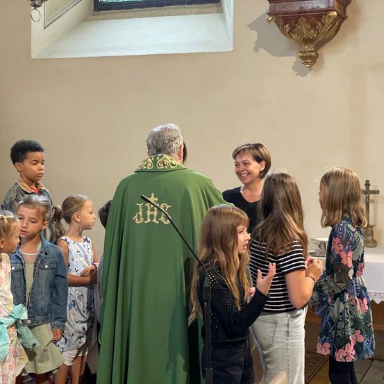 In einer Kirche steht ein Priester vor Kindern und lächelt. Sie sind in verschiedenen Outfits gekleidet, einige in Kleidern und andere in Jacken. Eine Frau mit kurzen Haaren steht neben ihnen.