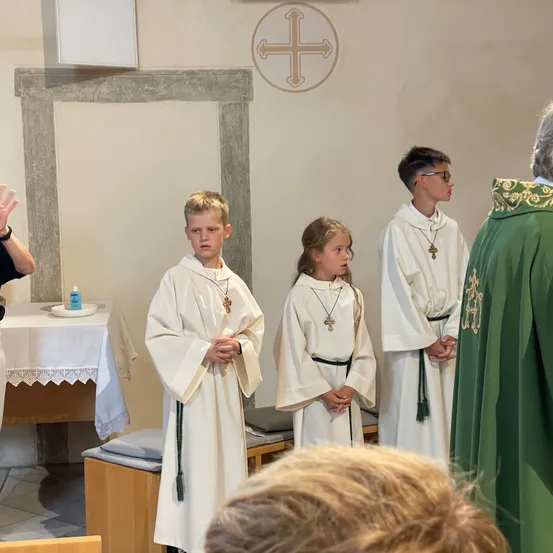Vier Kinder in weißen Gewändern, möglicherweise Messdiener, stehen in einer Kirche. Hinter ihnen steht ein Priester in grünen Gewändern. Links befindet sich ein Altar mit einem Tuch.