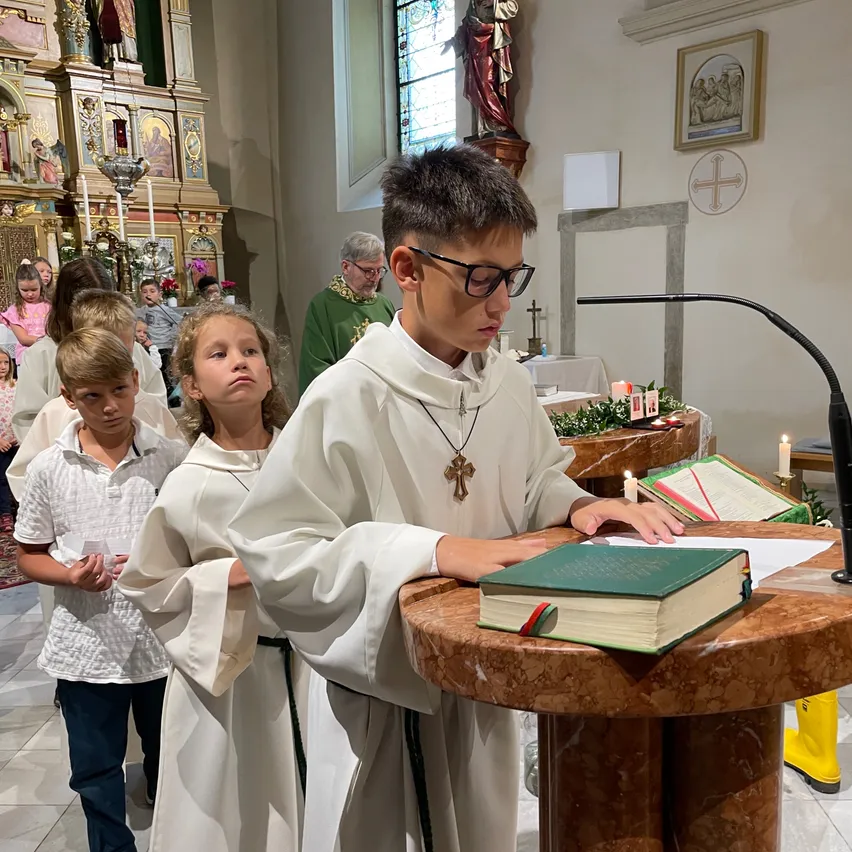 Ein junger Junge steht an einem Podest und liest aus einem Buch vor, umgeben von einer Gruppe von Kindern und Erwachsenen in einer Kirche.