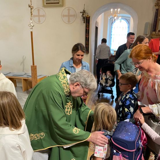 Ein Priester in grünen Gewändern kniet, um den Rucksack eines jungen Mädchens zu segnen, umgeben von Familien, in einer Kirche mit dekorativen Kreuzen und einer Kronleuchter.