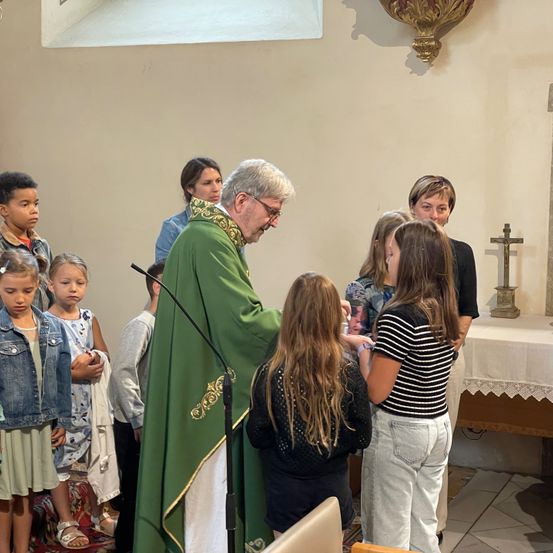 Ein Priester in grünen Gewändern führt eine Zeremonie in einer Kirche durch, umgeben von Kindern und Erwachsenen, von denen einer einen Segen erhält.
