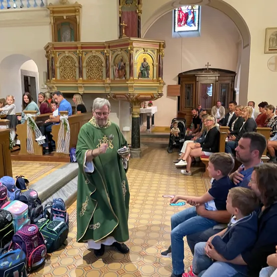 Ein Priester in einem grünen Gewand steht in einer Kirche, hält eine Schüssel und spricht zu einem sitzenden Publikum. Familien sitzen auf Bänken mit Rucksäcken um sie herum.