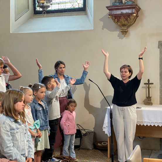 Eine Gruppe von Kindern und Erwachsenen steht in einer Kirche, einige mit erhobenen Händen. Eine Frau führt sie an und steht vor einem Mikrofon. Hinter ihr ist teilweise ein Tisch mit einem Kreuz zu sehen.