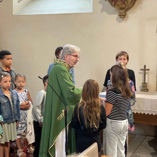 Ein Priester in einer grünen Robe steht an einem Pult in einer Kirche und spricht zu einer Gruppe von Kindern und Erwachsenen. Die Kinder, in verschiedenen Outfits gekleidet, hören aufmerksam zu. Ein Kreuz und eine Kerze stehen auf einem Tisch hinter dem Priester.