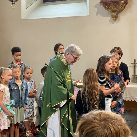 Ein Priester in grünen Gewändern wäscht den Füßen von kleinen Kindern in einer Kirche. Die Kinder stehen vor ihm und schauen nach oben. Hinter ihnen befindet sich ein Tisch und ein Kreuz auf dem Altar.