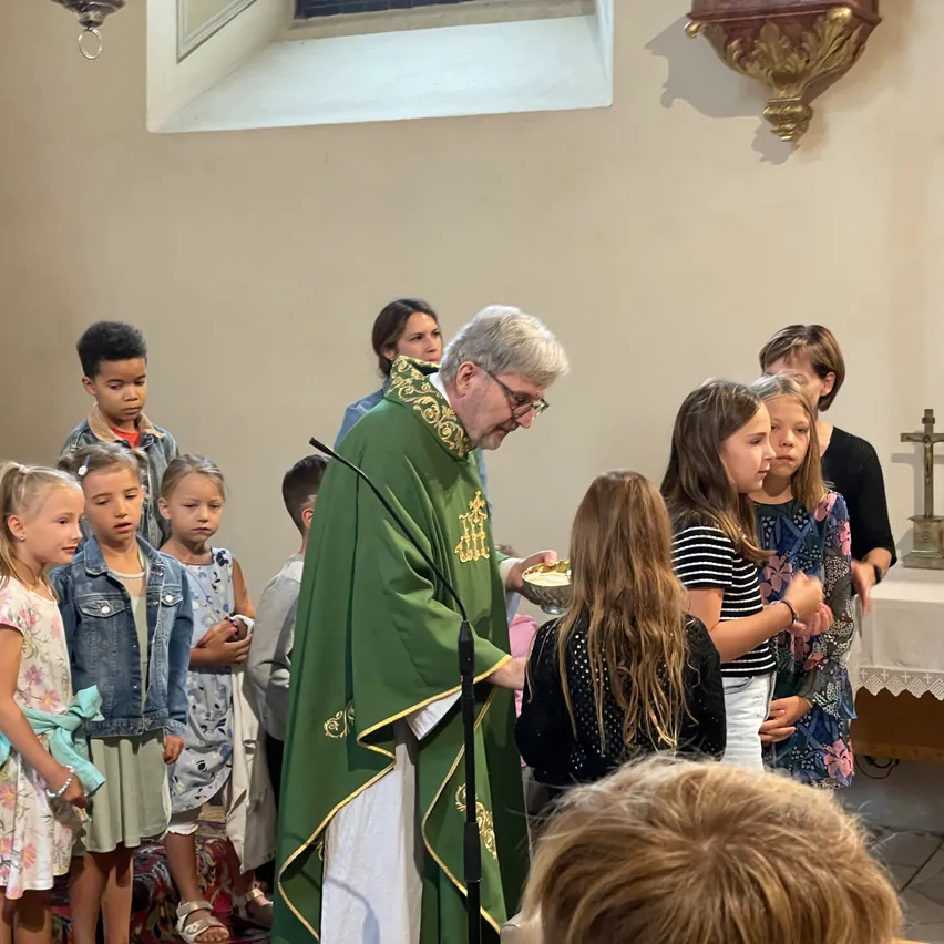 Ein Priester in grünen Gewändern wäscht den Füßen von kleinen Kindern in einer Kirche. Die Kinder stehen vor ihm und schauen nach oben. Hinter ihnen befindet sich ein Tisch und ein Kreuz auf dem Altar.